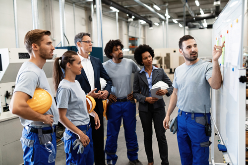 image of young employee wearing uniform presenting information to a group of colleagues, some in same uniform with hard hat, others in professional attire 