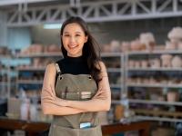 young female wearing apron smiles at the camera, shelves of pottery in the background