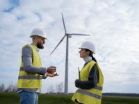 Male and young female wearing yellow vests and white hard hats talk with a windmill in the distance